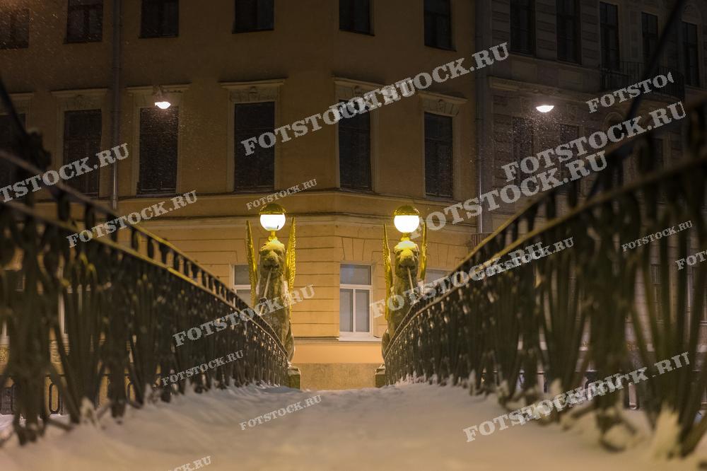 На фотографии изображен Банковский мост со знаменитыми грифонами зимней ночью. Мост украшен снежным покровом, а фонари освещают путь, создавая теплую атмосферу в холодную ночь. На заднем плане видны старинные здания города, добавляя ощущение уюта и старины.